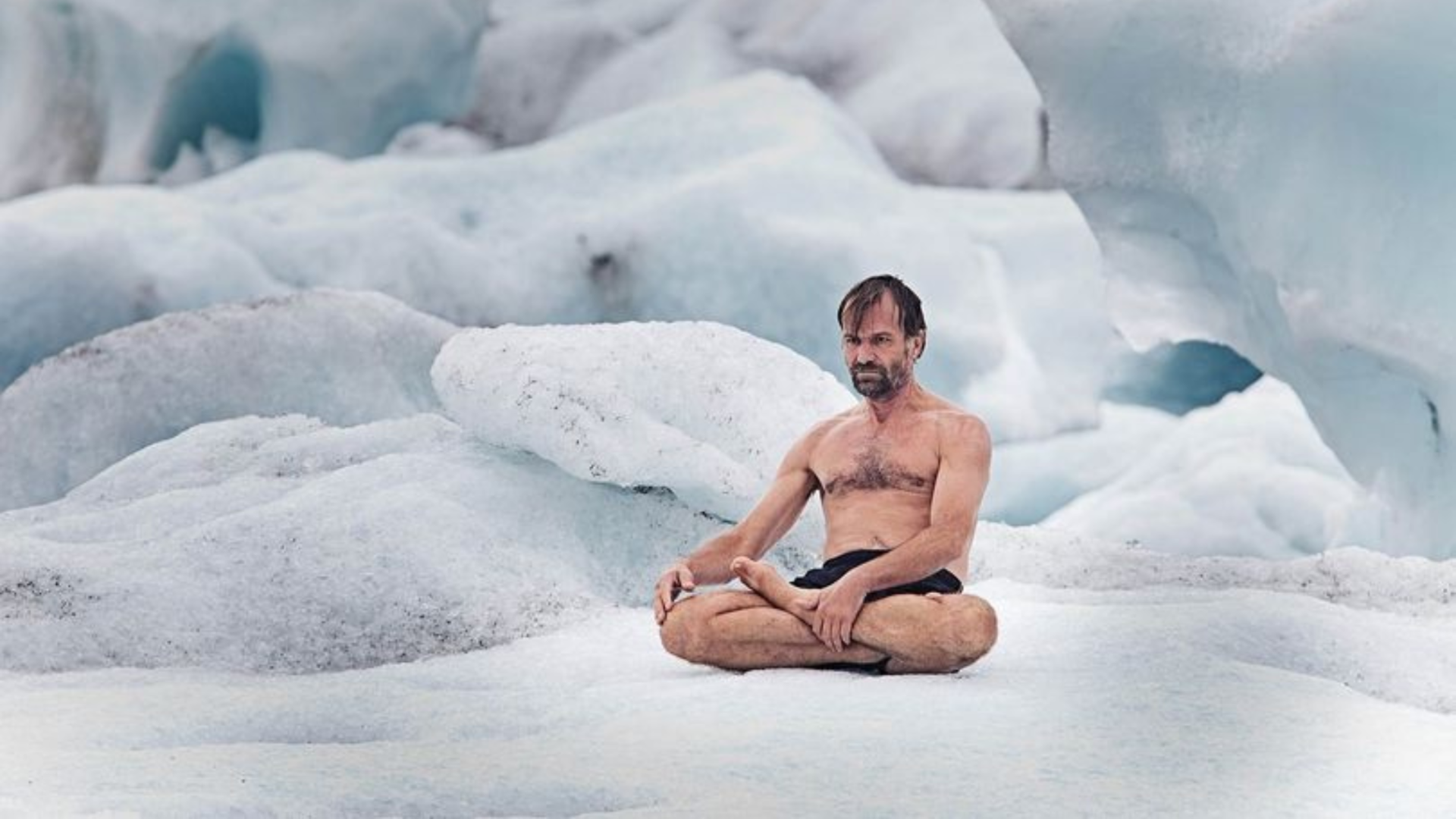 Wim Hof sedící v pozici tureckého sedu na sněhu, v pozadí ledové bloky a mlhavé modré hory.