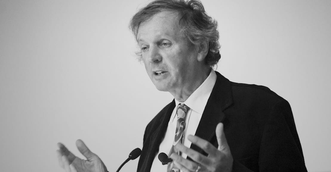 Černobílý portrét staršího muže, Ruperta Sheldrake, v saku s kravatou, mluví u mikrofonů a gestikuluje rukama.